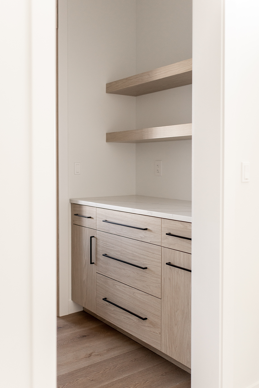 Walk-through pantry with natural wood finish cabinets, white marble countertop coffee station, and long dark square handles.