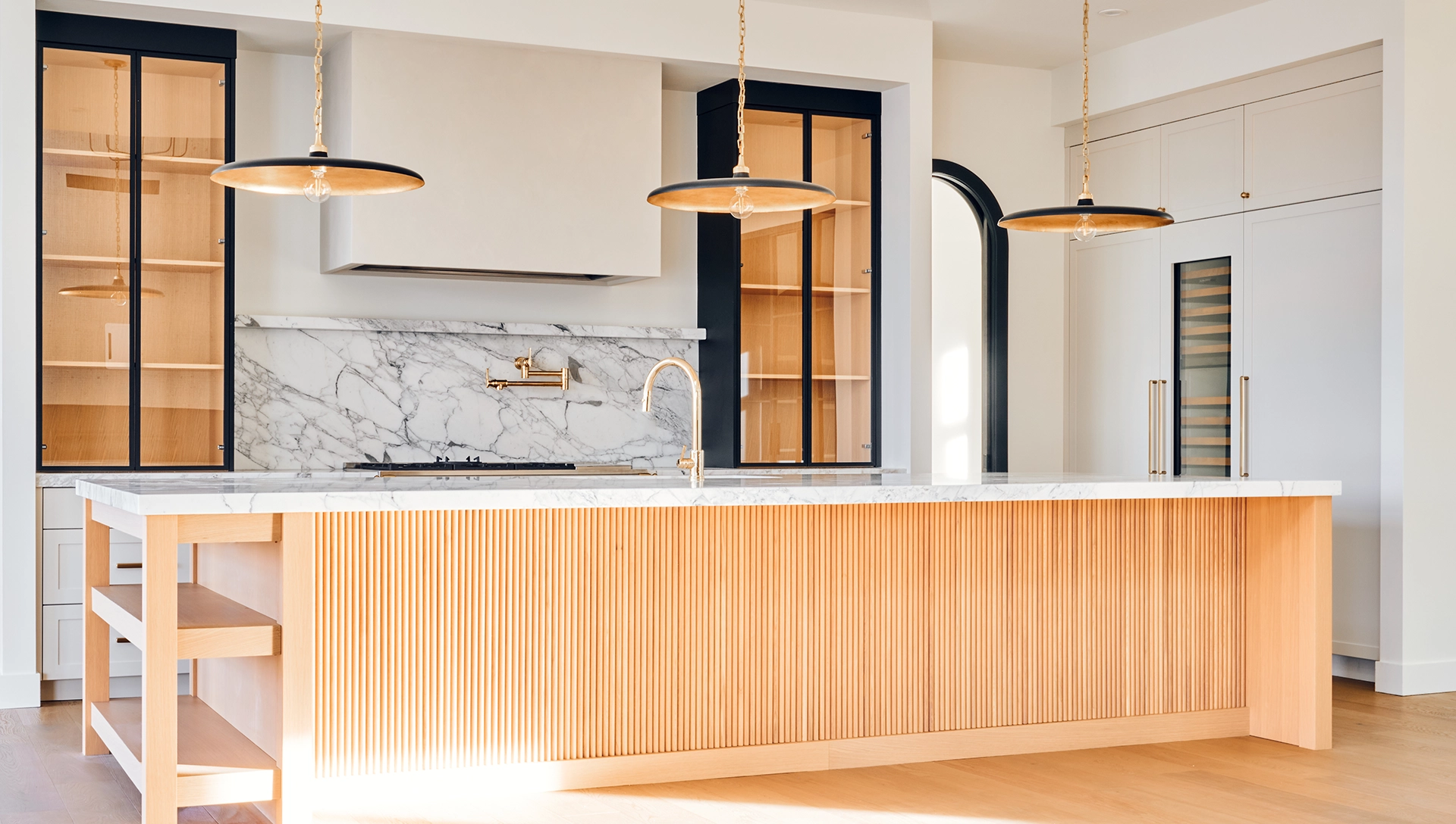 Custom kitchen with a fluted wood island, marble backsplash, black-framed glass cabinets and brass pendant lights
